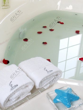 水中漂着玫瑰花瓣的浴盆旁摆放整齐的沐浴用品图片